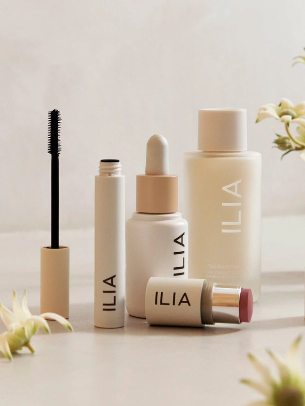 ILIA beauty products arranged on a neutral surface, including mascara, face milk, foundation, and a multi-stick with flowers in the background.