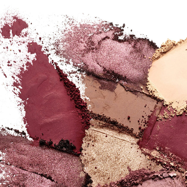 Crushed eyeshadow pigments in shades of burgundy, brown, and gold spread across a white background, showcasing texture and shimmer.