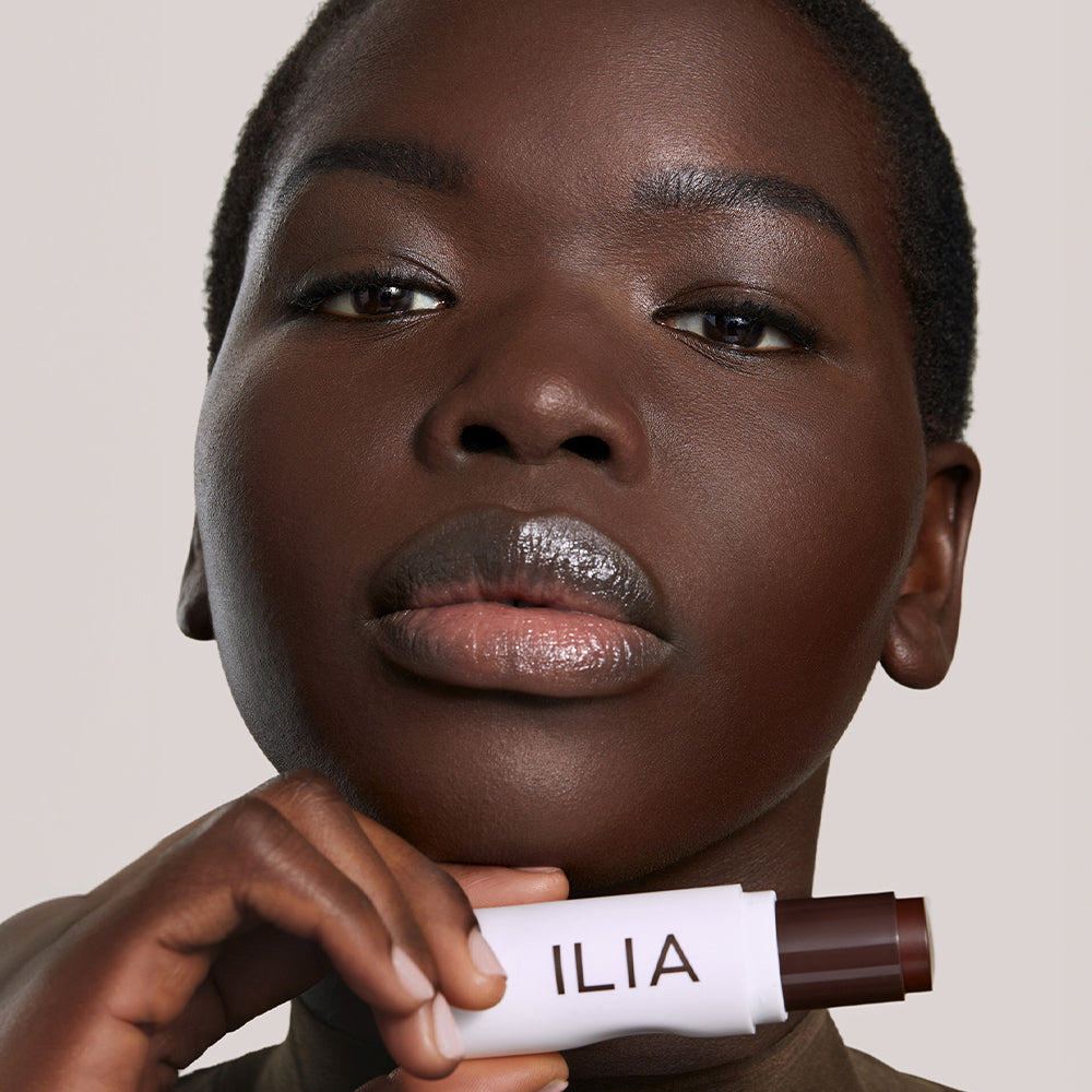 A model with a deeper skin tone holds an ILIA Complexion Stick, showcasing it's rich matte shade.