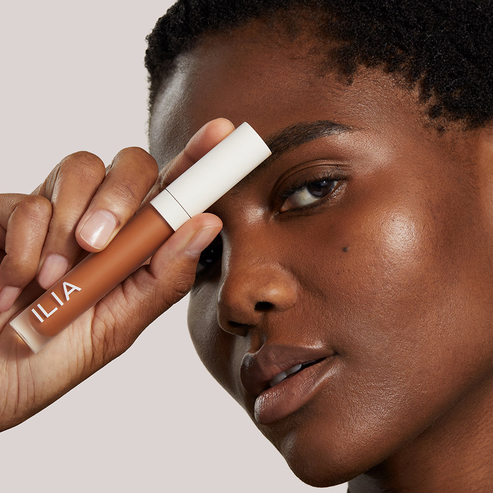 A model with radiant skin holds an ILIA concealer near her brow while gazing into the camera.