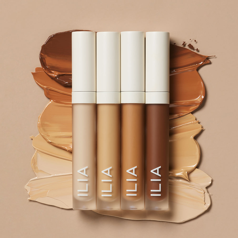 Four ILIA concealers stand upright against smeared swatches of different shades on a beige background.