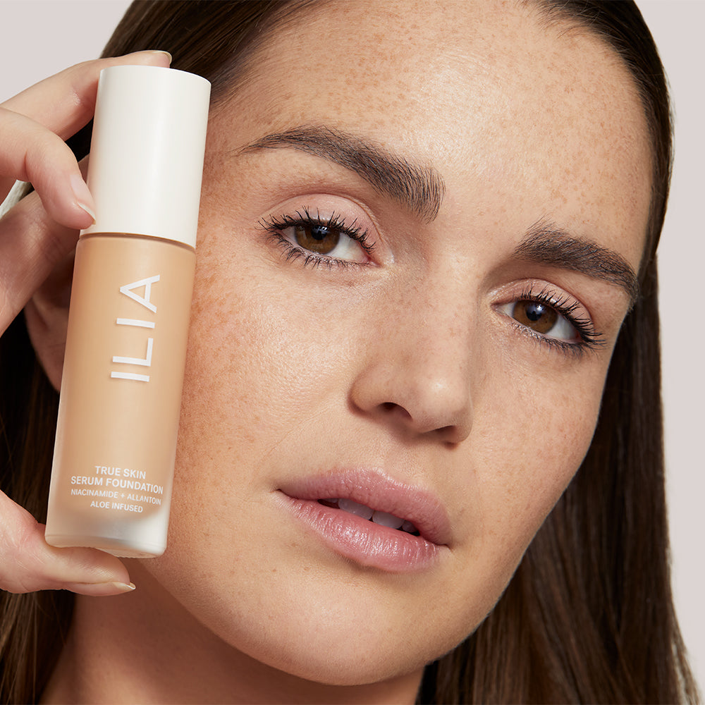 A model with freckles holds ILIA True Skin Serum Foundation near her face.