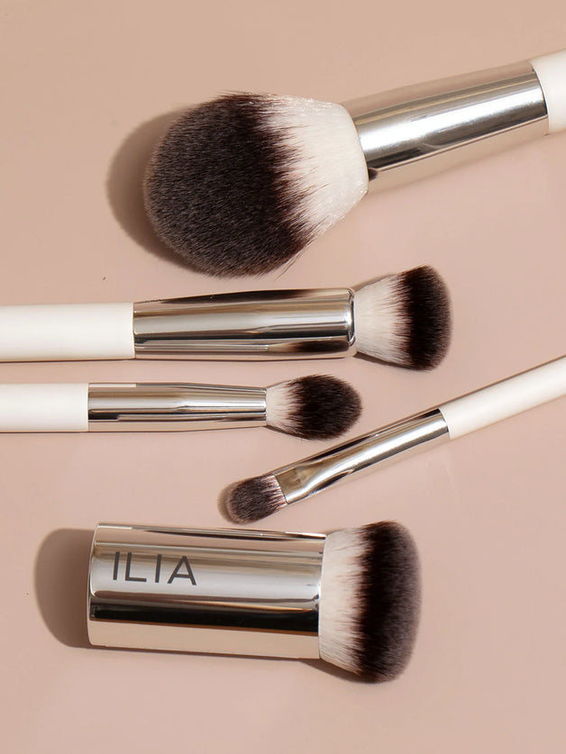 An array of ILIA brushes including fluffy, eyeshadow, dense brushes on a tan background.