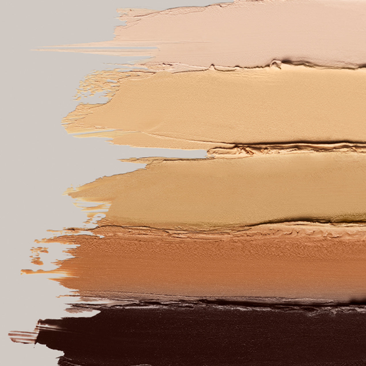 An array of ILIA's Skin Rewind Complexion Stick creamy swatches lined up against one another on a grey background.