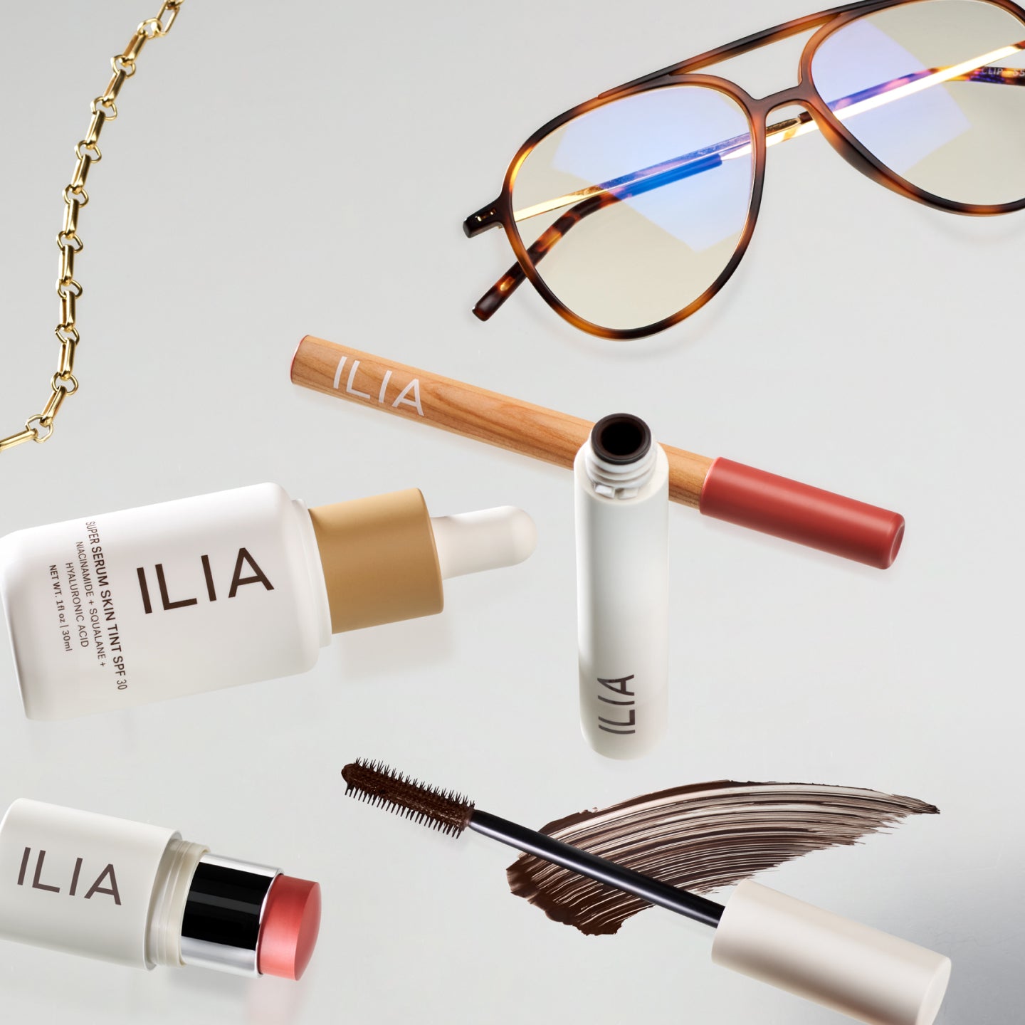 Assorted ILIA makeup items, sunglasses, and a gold chain displayed on a reflective surface.