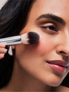 A person applies a pink blush to their cheek using ILIA Sculpting Brush.