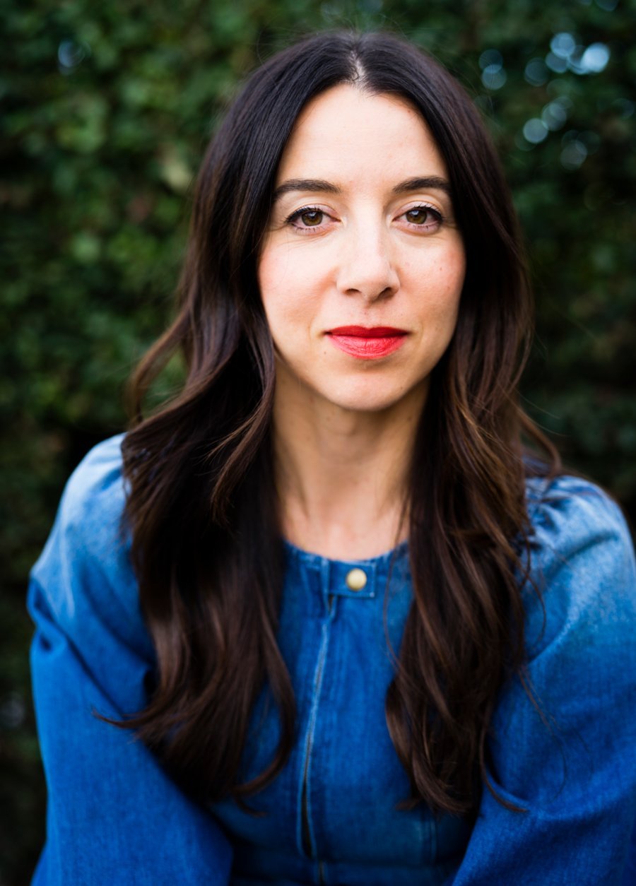 The founder of ILIA, Sasha, with a red  lipstick and denim top looks directly into the camera with a confident expression against a blurred greenery background.