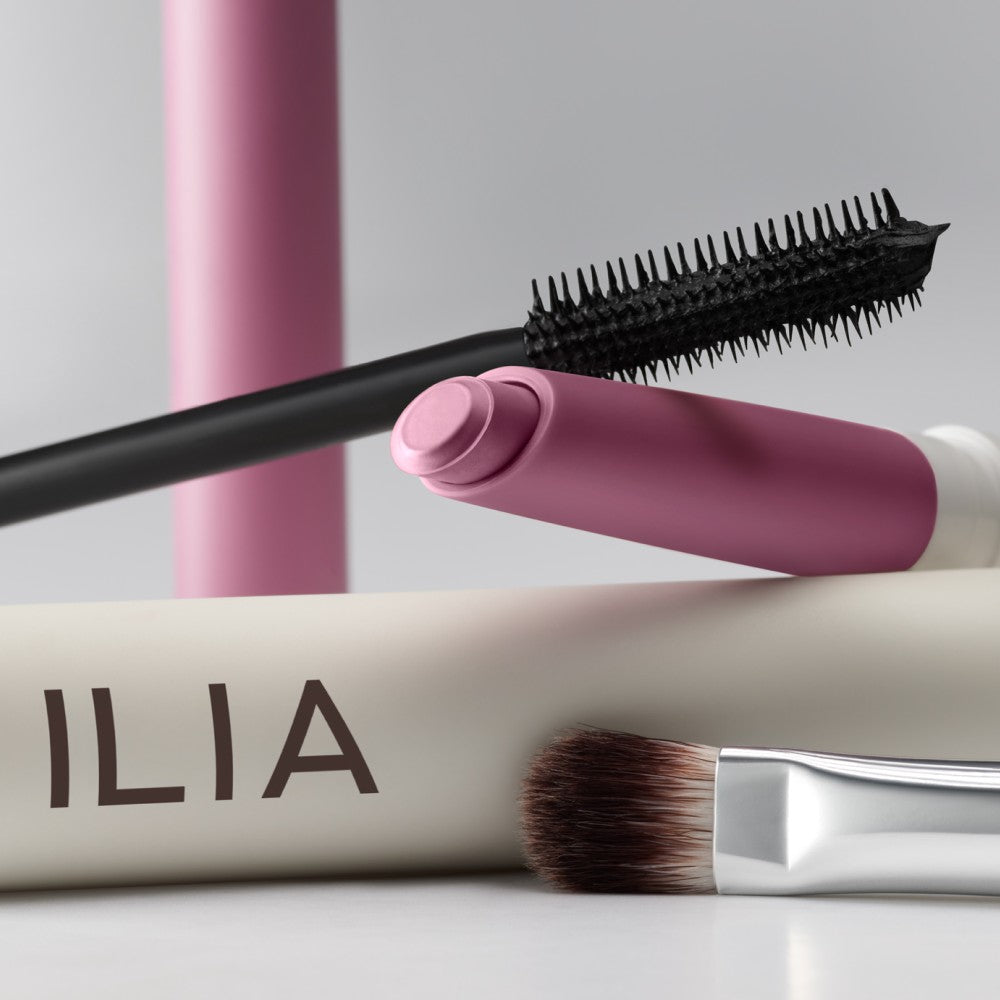 A variety of ILIA products including Eye Stylus, Mascara, and an eyeshadow brush, displayed diagonally.